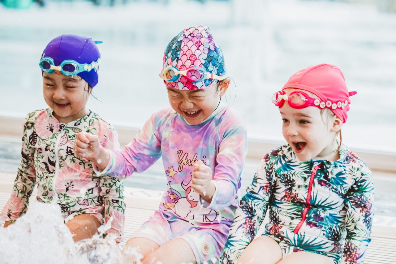 Three young girls in swim caps laughing and splashing on the pool edge at Pymble Swimming