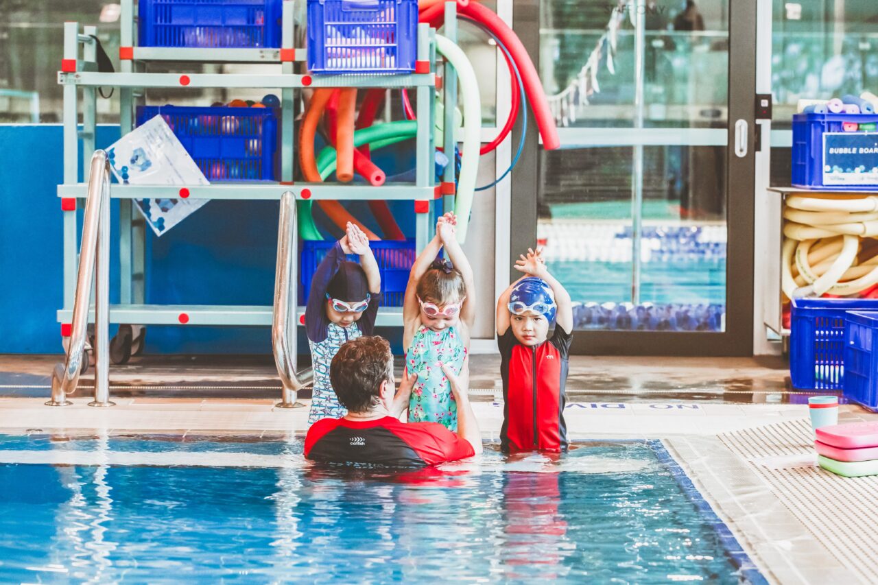 Pymble Swimming instructor leading three children through a stretch exercise on the pool steps